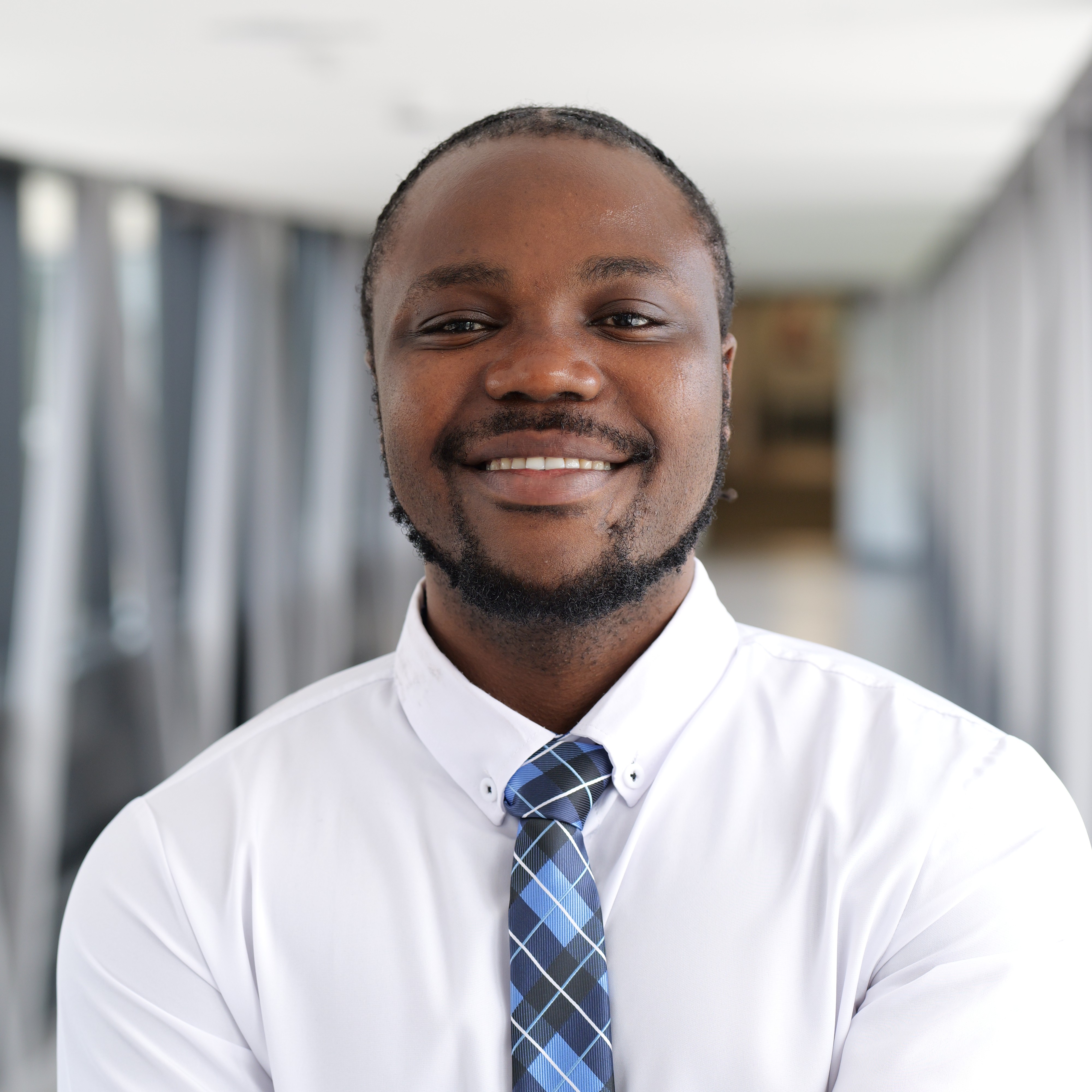 A picture of the current student president - Olakunle Stephen Adedoyin. He is a black man, in his mid 20s, smiling at the viewer, standing in a white shirt and and a blue tartan tie
