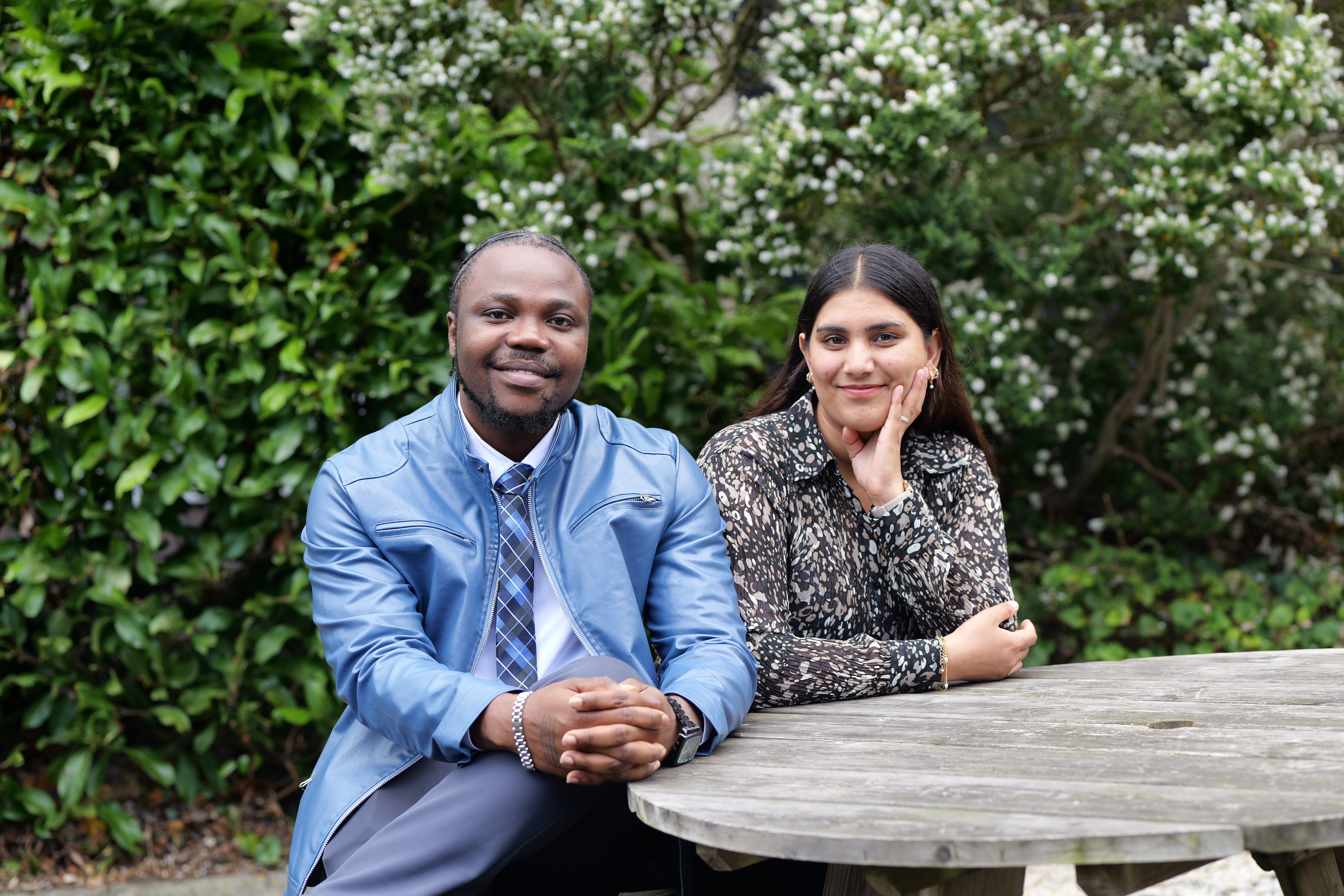 Your 2 student presidents sat at a table outdoors looking at the camera with flowering bushes behind them