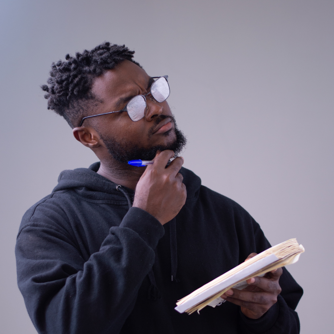 A black person with short hair and glasses looking thoughtful holding a pen and notepad