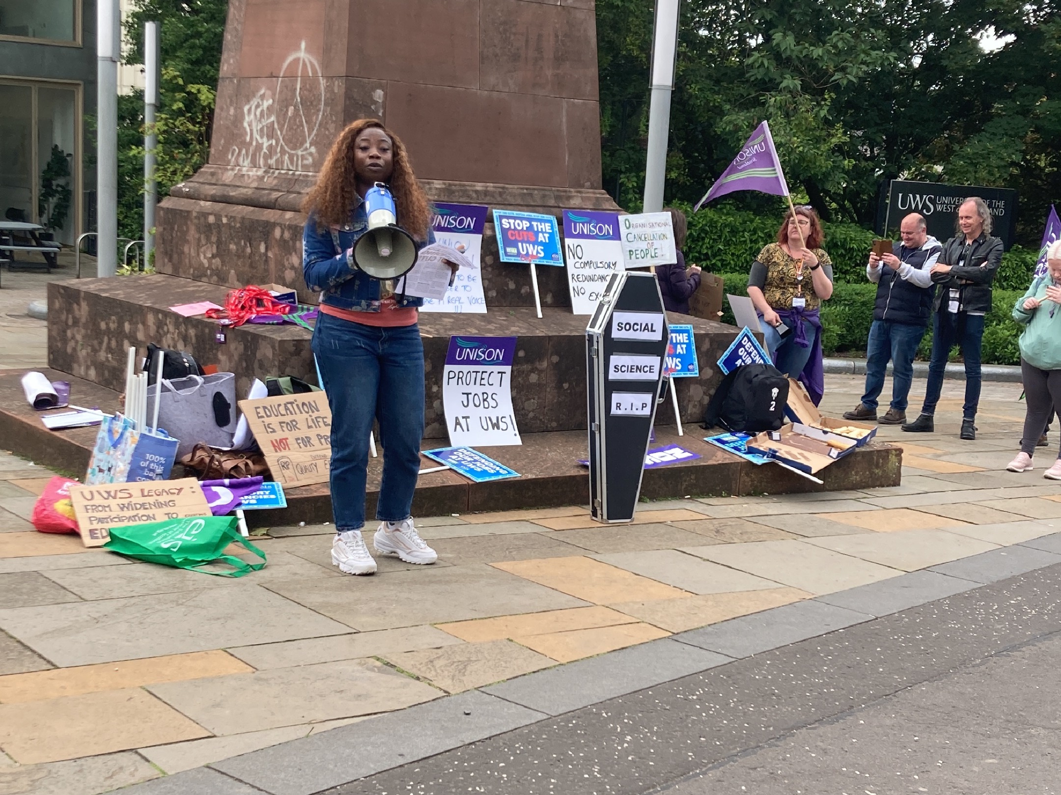 Omowaleola, the student president, speaking on a loudhailer standing in front or protest signs against the cuts